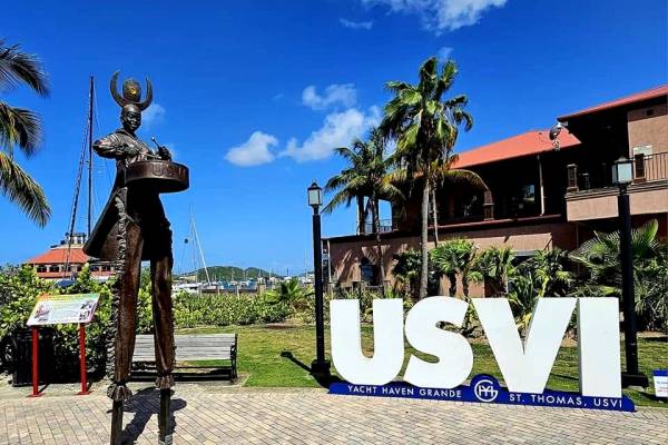 Large “USVI” sign at Yacht Haven Grande in St. Thomas, U.S. Virgin Islands, with a bronze waterfront sculpture, palm trees, marina buildings, and sailboat masts visible under a bright blue Caribbean sky.