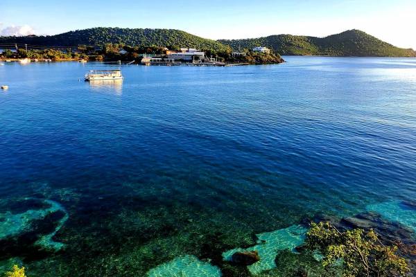 View of Water Bay in St. Thomas, U.S. Virgin Islands, featuring crystal-clear Caribbean waters, a small boat anchored offshore, lush green hills along the shoreline, and distant islands beneath a clear blue sky.