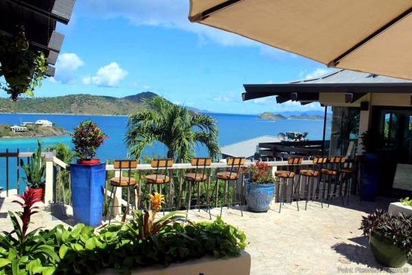 Outdoor bar and patio seating area at Lionfish Restaurant at Point Pleasant Resort in St. Thomas, U.S. Virgin Islands, featuring bar stools overlooking Water Bay, tropical landscaping, palm trees, and panoramic views of the Caribbean Sea and nearby islands under a sunny blue sky.