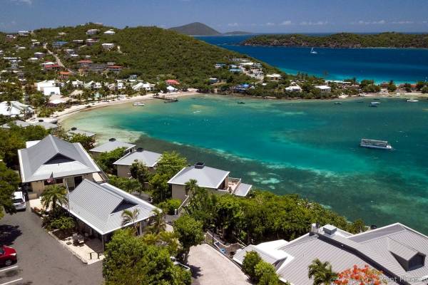 Aerial view of Point Pleasant Resort in St. Thomas, U.S. Virgin Islands, featuring hillside villas overlooking the turquoise waters of Water Bay, a sandy beach, anchored boats, lush tropical hills, and nearby Caribbean islands under a clear blue sky.