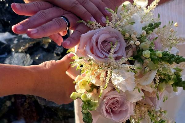 Close-up of two people wearing wedding bands stacking their hands together above a bouquet of pale pink roses and white flowers during a beach wedding ceremony.