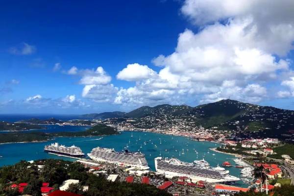 Elevated view of the WICO cruise ship dock in St. Thomas, U.S. Virgin Islands, with several large cruise ships moored in a turquoise harbor, sailboats scattered across the bay, and hillside neighborhoods under a bright blue sky with scattered clouds.
