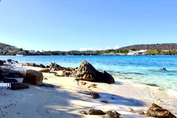 Sugar Beach at Point Pleasant Resort in St. Thomas, U.S. Virgin Islands, featuring a sandy shoreline with lounge chairs, rocky coastal formations, calm turquoise Caribbean water, and scenic island views across the bay under a clear blue sky.