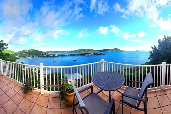 Panoramic terrace view at Point Pleasant Resort in St. Thomas, U.S. Virgin Islands, featuring outdoor seating overlooking Water Bay, lush green hills, anchored boats, and surrounding Caribbean islands beneath a bright blue sky with scattered clouds.