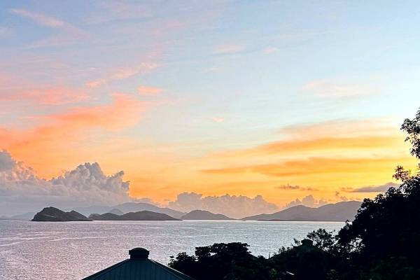 Sunrise view over the Caribbean Sea from Point Pleasant Resort in St. Thomas, U.S. Virgin Islands, featuring colorful orange and pink skies, calm ocean waters, silhouetted islands on the horizon, and lush tropical vegetation in the foreground.