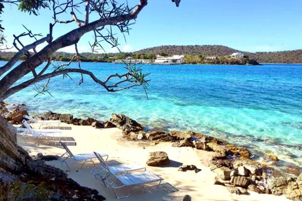 Sugar Beach at Point Pleasant Resort in St. Thomas, U.S. Virgin Islands, featuring lounge chairs on a sandy shoreline, rocky outcroppings, crystal-clear turquoise water, and tropical tree branches framing the scenic Caribbean bay.