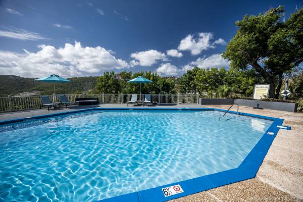 Upper pool at Point Pleasant Resort in St. Thomas, U.S. Virgin Islands, featuring a sparkling outdoor swimming pool surrounded by lounge chairs and blue umbrellas, with lush tropical greenery, rolling hills, and a bright blue sky filled with white clouds.