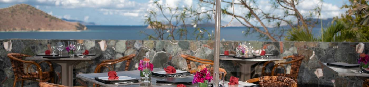 Nicely set tables on a patio with Water Bay and various cays of the Virgin Islands in the background on a sunny day.